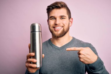 Sarışın, sakallı, mavi gözlü, termosu pembe arka planda tutan genç bir adam.