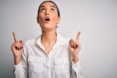 Genç, güzel, esmer bir kadın, izole edilmiş beyaz arka plan üzerinde gündelik bir gömlek giyiyor. Şaşırmış ve şaşırmış bir şekilde yukarı bakıyor ve parmaklarıyla işaret ediyor ve kollarını kaldırıyor..