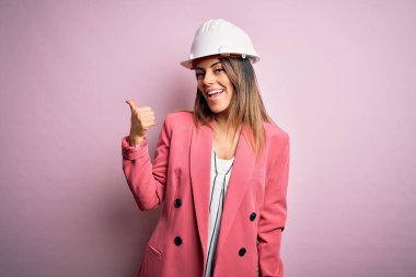 Genç, güzel esmer mimar kadın pembe arka planda güvenlik kaskı takıyor. Gülümsüyor, gülümsüyor ve başparmağıyla yana bakıyor..