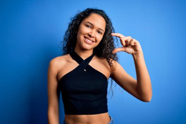 Kıvırcık saçlı genç bir kadın mavi arka planda dikilmiş tişört giyiyor gülümsüyor ve el hareketleriyle küçük beden işareti yapıyor ve kameraya bakıyor. Ölçü kavramı.