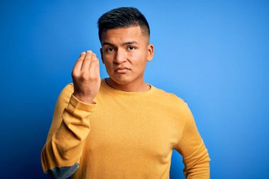 Sarı günlük süveter giyen genç, yakışıklı latin adam izole edilmiş mavi arka planda İtalyan jesti yapıyor ve parmakları kendinden emin.