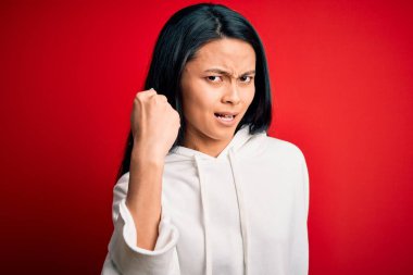 Genç ve güzel Çinli sporcu kadın izole edilmiş kırmızı arka plan üzerine kazak giyiyor kızgın ve kızgın yumruğunu kaldırıyor ve öfkeyle bağırıyor. Öfke ve agresif kavram.