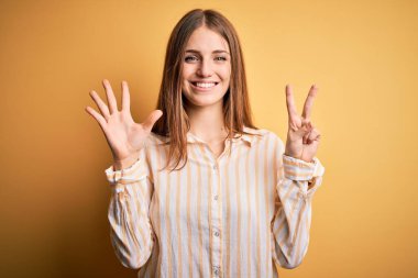 Genç, güzel, kızıl saçlı, günlük çizgili gömlek giyen, izole edilmiş sarı arka planda, yedinci parmaklarıyla işaret eden, kendinden emin ve mutlu bir şekilde gülümseyen bir kadın..