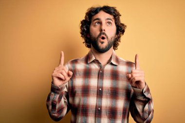 Sarı arka planda dikilen sıradan tişörtlü genç yakışıklı adam şaşırdı ve şaşırdı. Parmaklarıyla işaret etti ve kollarını kaldırdı..