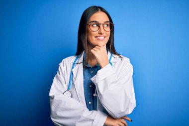 Genç, güzel doktor kadın steteskop takıyor ve mavi arka planda gözlük takıyor, elleri çenesinde soru sorarken, düşünceli bir ifade takınıyor. Düşünceli bir yüzle gülümsüyor. Şüphe kavramı.