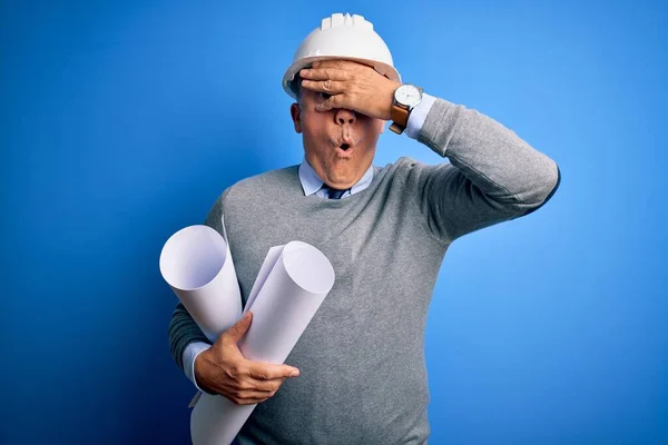 Middle age handsome grey-haired architect man wearing safety helmet holding blueprints peeking in shock covering face and eyes with hand, looking through fingers with embarrassed expression.