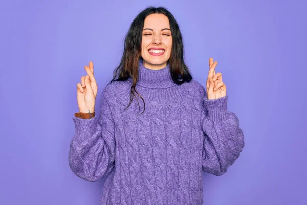 Young beautiful woman wearing casual turtleneck sweater standing over purple background gesturing finger crossed smiling with hope and eyes closed. Luck and superstitious concept.