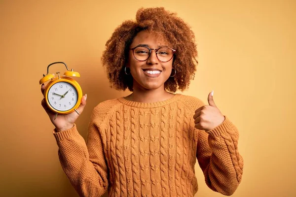 Young beautiful African American afro woman with curly hair holding vintage alarm clock happy with big smile doing ok sign, thumb up with fingers, excellent sign