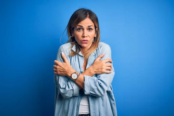 Middle age beautiful woman wearing casual shirt standing over isolated blue background shaking and freezing for winter cold with sad and shock expression on face