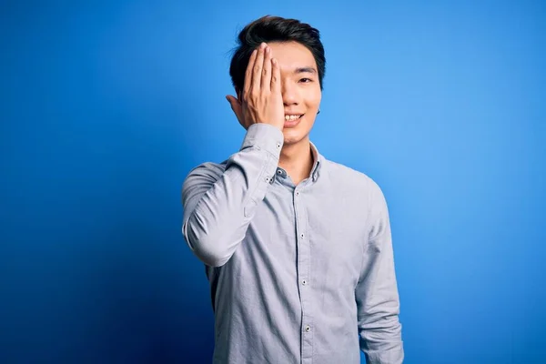 若いですハンサムな中国人男性身に着けているカジュアルシャツ上に立って隔離された青の背景カバー1目で手 顔に自信を持って笑顔と驚きの感情 — ストック写真