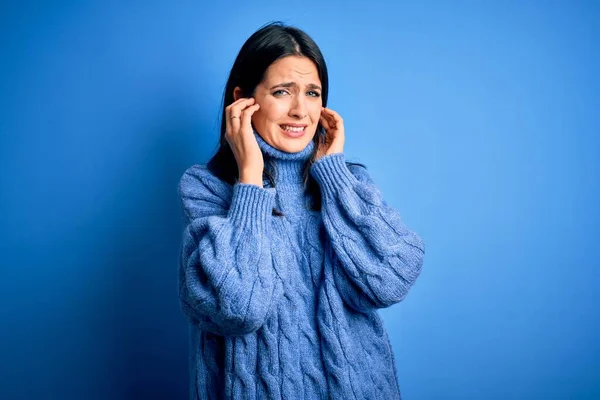 青い目をした若いブルネットの女性は 大きな音楽のノイズのためにいらいらした表情で指で耳を覆うカジュアルなタートルネックセーターを着ています ろう者の概念 — ストック写真