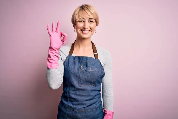 年轻的金发清洁女人 留着围裙 戴着粉色背景的手套 微笑着 手指手画脚地签了名 成功表达 — 图库照片