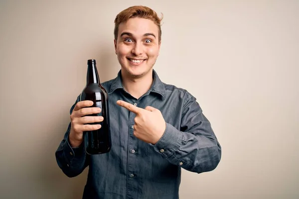 年轻英俊的红头发男人在与世隔绝的白色背景下喝着一瓶啤酒 手指手画脚地笑了笑 — 图库照片