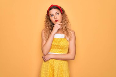 Sarı arka planda dikey duran mavi gözlü güzel sarışın kadın. Elleri çenesinde, soru sormayı, düşünceli ifadeyi düşünüyor. Düşünceli bir yüzle gülümsüyor. Şüphe kavramı.