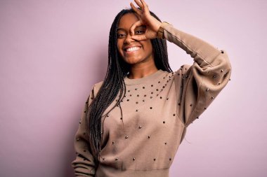 Pembe izole edilmiş arka planda moda süveteri giyen Afro-Amerikalı genç bir kadın el hareketleriyle gülümsüyor ve mutlu yüzle parmaklarının arasından bakıyor..