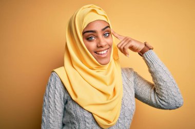 Genç ve güzel Afro-Amerikalı kız izole edilmiş sarı arka planda Müslüman başörtüsü takıyor. Tek parmağıyla başını işaret ediyor. Harika bir fikir ya da düşünce, iyi bir hafıza.
