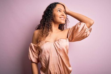 Kıvırcık saçlı, günlük tişörtünü giymiş pembe arka planda duran, kendine güvenen, el hareketleriyle saçlarına dokunan, çekici ve modaya uygun pozlar veren güzel bir kadın.