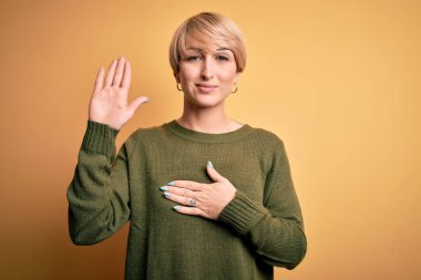 Modern kısa saçlı, sarı arka planda rahat süveter giyen, elleri göğsünde ve avucunda açık yemin eden, sadakat yemini eden genç sarışın bir kadın.