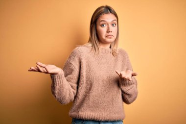 Sarı arka plan üzerinde kahverengi süveter giyen genç, sarışın bir kadın. Kolları ve elleri havada, kafası karışmış bir yüz ifadesi. Şüphe kavramı.