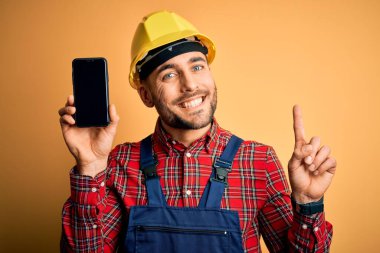 Güvenlik kaskı takan genç müteahhit sarı arka planda akıllı telefon ekranı gösteriyor.