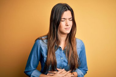 Genç ve güzel bir kız günlük kot gömleği giyerek izole edilmiş sarı arka planda duruyor. Midenin üstünde el ele. Çünkü hazımsızlık, acı verici bir hastalık, kendini iyi hissetmiyor. Ache kavramı.