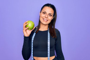 Güzel esmer sporcu kadın, mezura kullanarak mor arka planda elma meyvesi yiyor. Olumlu ve mutlu görünüyor. Kendine güvenen bir gülümsemeyle dişlerini gösteriyor.