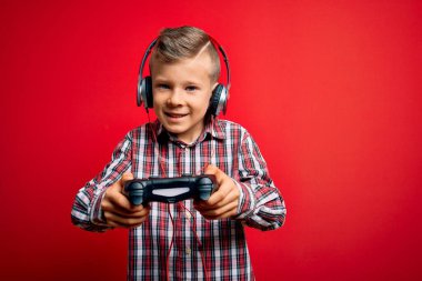 Sevimli sarışın oyuncu çocuk mutlu ve kendinden emin gülümsüyor. Yüzünde tebessümle dikilip, izole edilmiş kırmızı arkaplan üzerinde gamepad ve kulaklık kullanarak video oyunu oynuyorum.