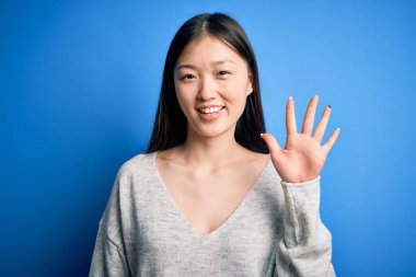 Genç ve güzel Asyalı bir kadın günlük süveter giyiyor mavi izole edilmiş arka plan üzerinde duruyor ve parmaklarını beş numarayı gösterirken kendinden emin ve mutlu gülümsüyor..