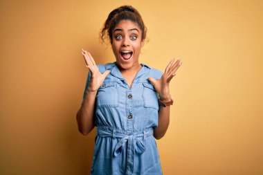 Sarı arka planda dikilen kot elbiseli güzel Afro-Amerikan kızı çılgın ve başarıyı kollarını kaldırıp heyecanla haykırarak kutluyor. Kazanan konsept