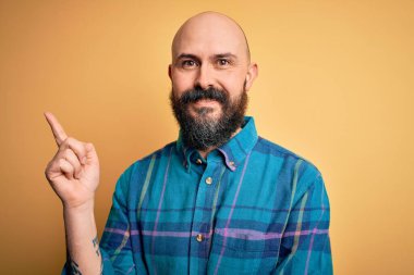 Sarışın, sakallı, sıradan tişörtlü, yalıtılmış sarı arka planda duran, yüzünde kocaman bir gülümseme olan, eli ve parmağı kameraya bakan kel bir adam..