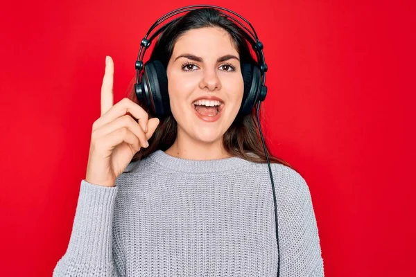 現代のヘッドフォンを身に着けている若い美しい女の子は 成功したアイデアで指を指す赤い背景の上に音楽に耳を傾けます 興奮し 第一番 — ストック写真