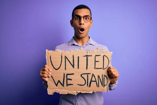 African american activist man asking for union holding banner with united stand message scared in shock with a surprise face, afraid and excited with fear expression