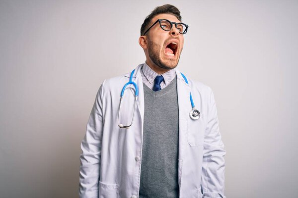 Young doctor man with blue eyes wearing medical coat and stethoscope over isolated background angry and mad screaming frustrated and furious, shouting with anger. Rage and aggressive concept.