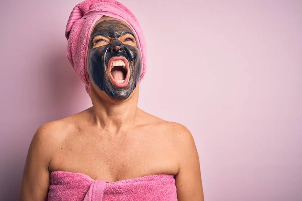 Middle age brunette woman wearing beauty black face mask over isolated pink background angry and mad screaming frustrated and furious, shouting with anger. Rage and aggressive concept.