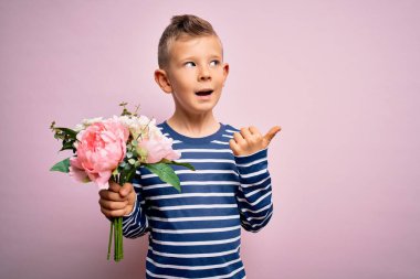 Küçük beyaz bir çocuk pembe arka planda romantik ve şirin bir buket çiçek tutuyor ve gülümseyen bir gülümsemeyle baş parmağıyla işaret ediyor.