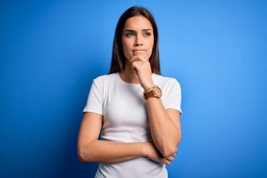 Genç, güzel, esmer, beyaz tişörtlü bir kadın. Mavi arka planda duruyor. Elleri çenesinde soru sorarken, düşünceli bir yüz ifadesi var. Düşünceli bir yüzle gülümsüyor. Şüphe kavramı.