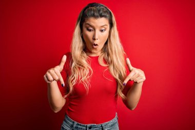 Genç ve güzel sarışın bir kadın, günlük tişört giyiyor, kırmızı arka plan üzerinde duruyor, parmaklarını göstererek reklamını yapıyor, şaşırmış yüzünü ve ağzını açıyor.