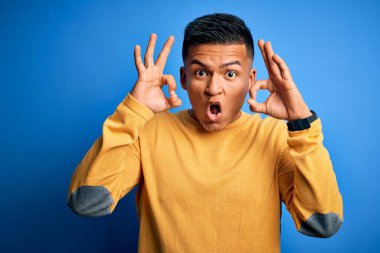 İzole edilmiş mavi arka plan üzerinde sarı günlük süveter giyen genç, yakışıklı latin adam parmaklarıyla onay sembolü yaparken şaşırmış ve şok olmuş görünüyor. Çılgın ifade