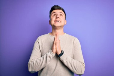 Mor izole edilmiş arka plan üzerine günlük süveter giyen yakışıklı beyaz bir adam elleri bağlı yalvarıyor ve dua ediyor. Yüzünde umut ifadesi var. Çok duygusal ve endişeli. Yalvarıyorum..