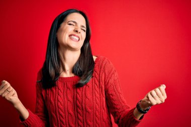 Mavi gözlü genç esmer kadın izole edilmiş kırmızı arka plan üzerine gündelik süveter giyiyor. Çok mutlu ve heyecanlı. Kolları havada, gülümseyen ve başarı için çığlık atan kazanan bir jest yapıyor. Kutlama konsepti.