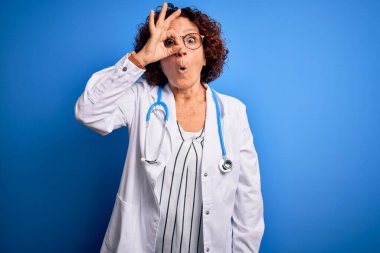 Mavi arka plan üzerinde steteskop ve ceket giyen orta yaşlı kıvırcık saçlı kadın şaşırmış yüz ifadesiyle şaşırmış, parmaklarından bakan gözler. İnançsız ifade.