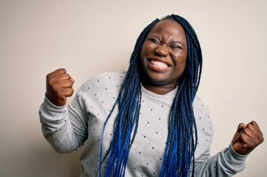 Beyaz arka plan üzerine günlük kazak giyen Afrikalı Amerikalı artı cüsseli kadın çok mutlu ve heyecanlı bir şekilde kolları havada, gülümseyerek ve başarı için çığlık atarak kazanan jestler yapıyor. Kutlama konsepti.