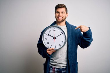 Sakallı ve mavi gözlü genç bir adam pijama giyiyor. Elinde kocaman bir saat var.