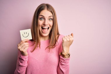Genç, güzel, kızıl saçlı bir kadın elinde gülümseme kağıdıyla emoji mesajı atıyor. Gururla bağırıyor, zaferi ve başarıyı kutluyor. Çok heyecanlı, coşkulu bir duygu.