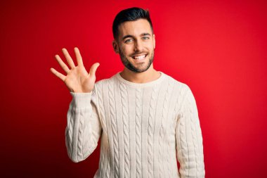 İzole edilmiş kırmızı arka plan üzerinde duran, rahat beyaz süveter giyen genç ve beş numaralı parmaklarıyla işaret eden kendinden emin ve mutlu gülümseyen bir adam..