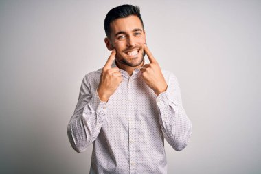 Zarif gömlekli genç adam izole edilmiş beyaz arka planda duruyor ağzı açık gülümsüyor, parmakları işaret ediyor ve zorla neşeli bir gülümseme sergiliyor.