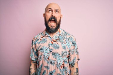Sakallı ve dövmeli yakışıklı, pembe arka planda sıradan bir çiçek gömleği giyen sinirli, sinirli ve öfkeli bir adam. Öfkeyle bağırıyor. Öfke ve agresif kavram.
