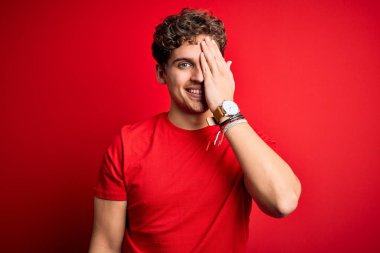 Sarışın, kıvırcık saçlı, kırmızı arka planda sıradan tişörtler giyen, bir gözünü kapayan, yüzünde kendine güvenen bir gülümseme ve sürpriz bir duygu olan genç bir adam..