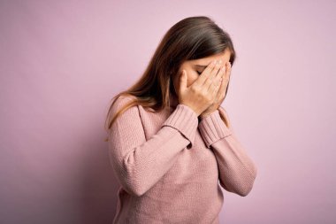 Güzel genç bir kadın pembe izole edilmiş arka plan üzerine balıkçı yaka kazak giyiyor ve ağlarken suratını ellerle kapatıyor. Depresyon kavramı.