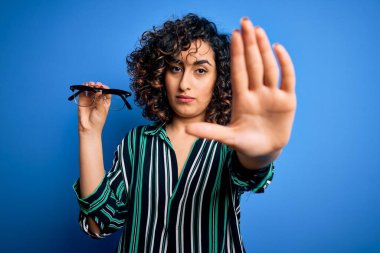 Güzel, optik Arap bir kadın, elinde vizyon gözlüğü, mavi arka planda, açık elle, dur işareti, ciddi ve kendinden emin bir ifade, savunma hareketi yapıyor.